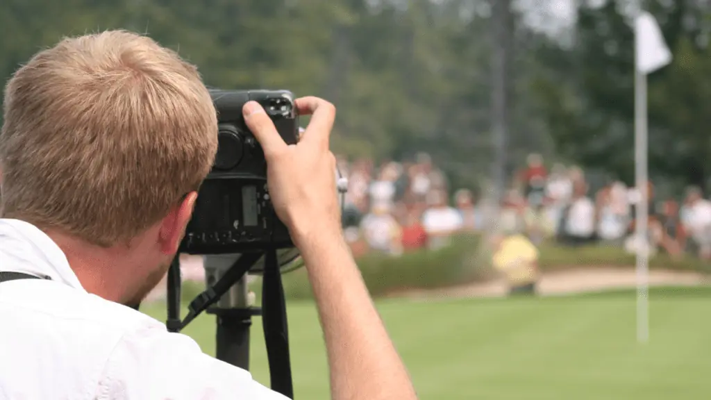 Photo of photographer and crowds around the green as golfer hits his shot out of the bunker. 