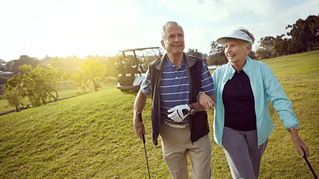 Senior couple golfing and holding arms as they walk toward the green.