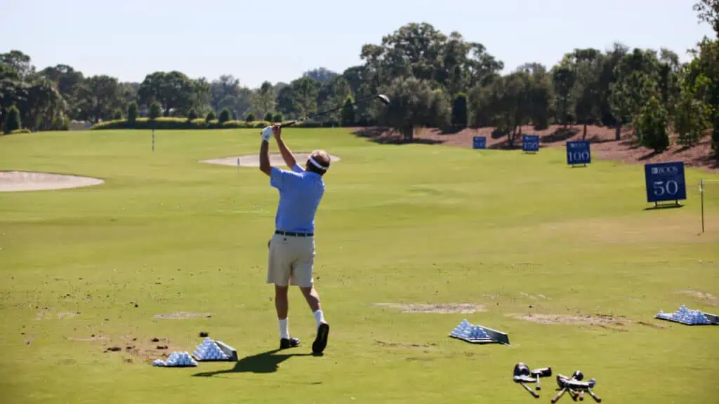 older man, senior golfer testing golf clubs hitting a fairway wood with a pile of other golf clubs on the ground at the driving range